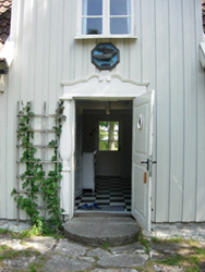 En öppen dörr, en ros klättrar bredvid. I hallen innanför ses ett svart-och-vitrutigt golv och längst in en fönsterdörr