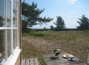 Ett enkelt bord, anar stugans fasad till vänster, framför en liten strandäng och i fonden anas havet